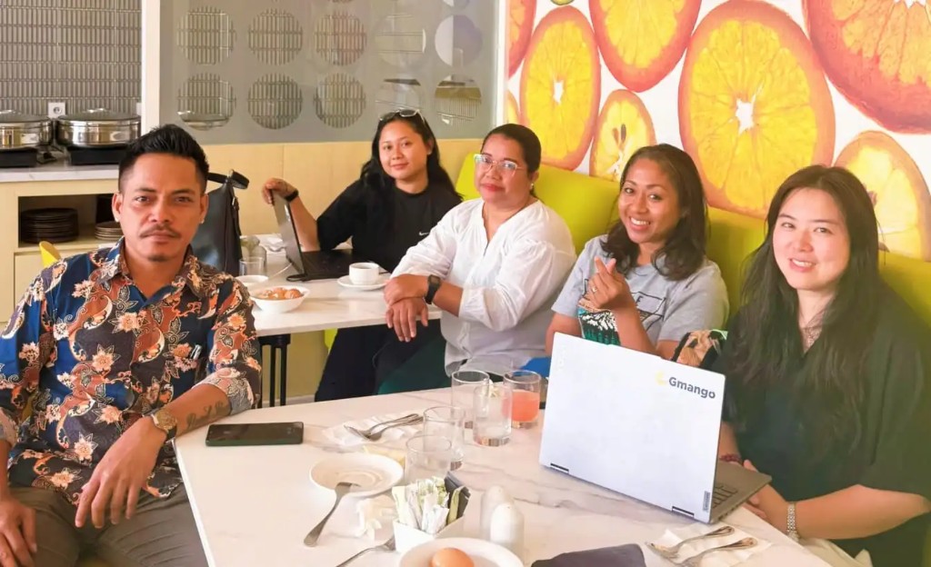 Five people at a café table, with a Gmango laptop and an orange-slice wall behind them